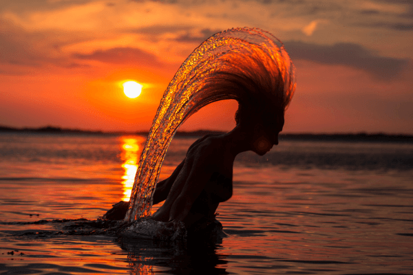 Pessoa jogando os cabelos para trás na água durante o pôr do sol, criando um arco de gotas iluminado pela luz dourada do entardecer.